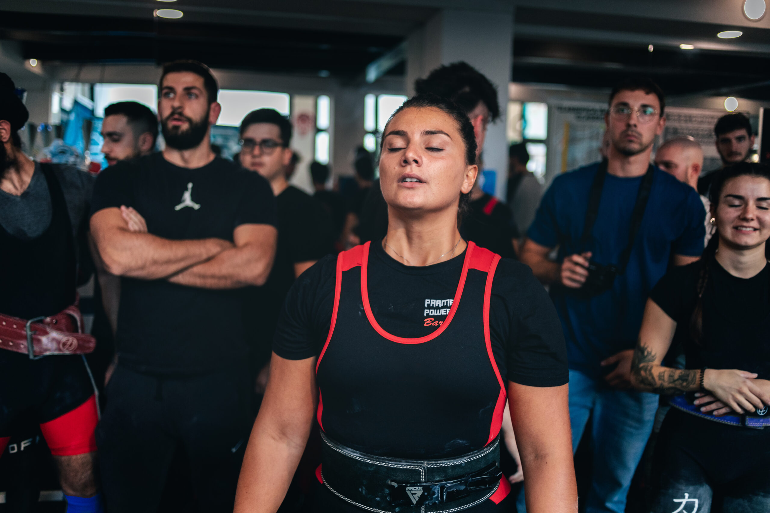 Una mujer concentrada antes de un concurso de powerlifting, rodeada de personas que observan a los otros participantes.