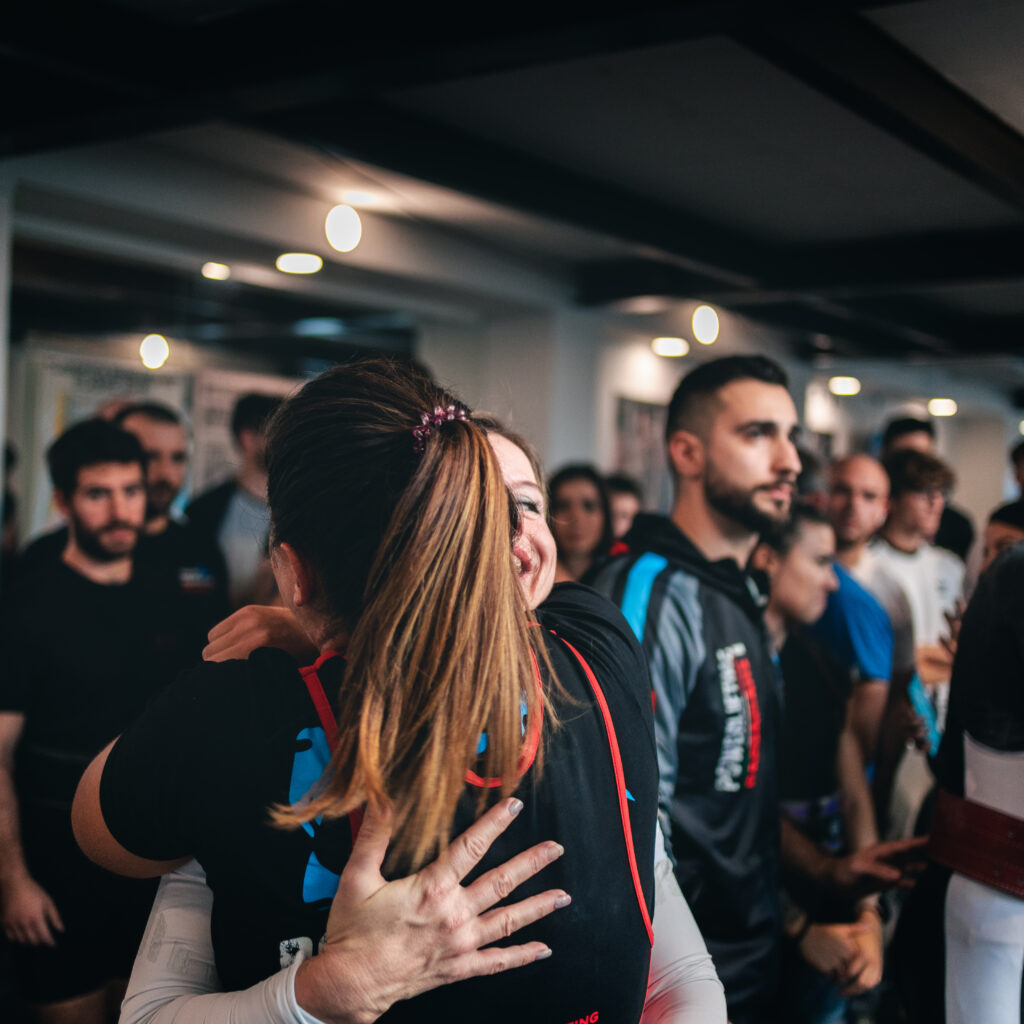 En primer plano, una mujer abraza a su entrenadora durante una competencia, con otras personas del evento al fondo.