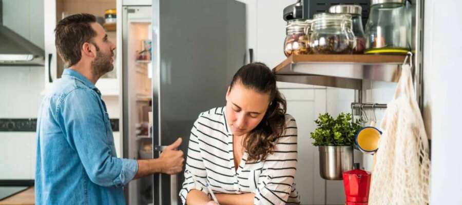 Hombre mirando en el refrigerador mientras una mujer escribe una lista de compras en la encimera de la cocina