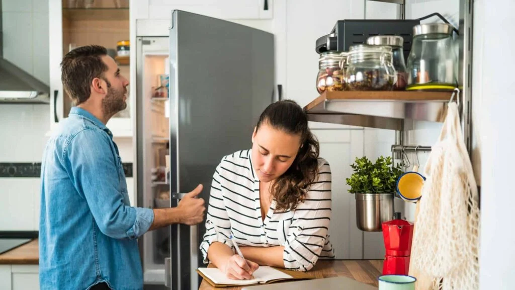 Hombre mirando en el refrigerador mientras una mujer escribe una lista de compras en la encimera de la cocina