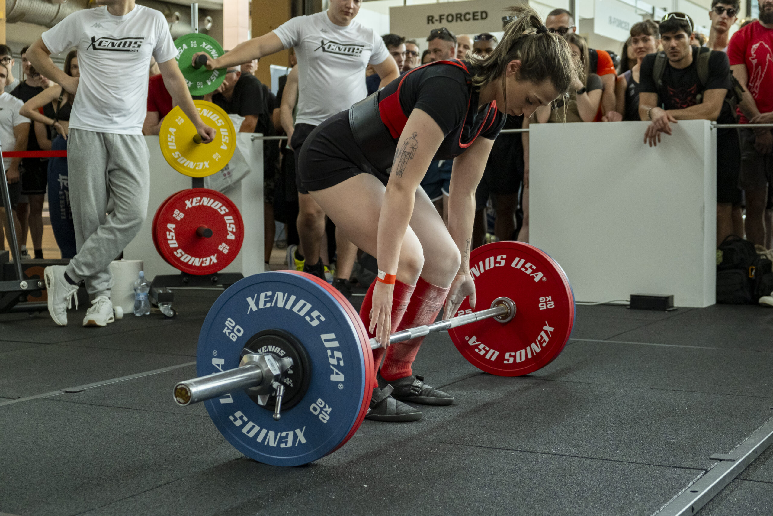Una mujer se prepara para realizar un levantamiento de peso durante una competencia de powerlifting. En el fondo, un juez y una audiencia observan el evento.